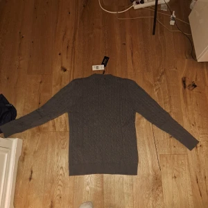 Grå stickad tröja Polo Ralph Lauren - Snygg grå stickad tröja från Polo Ralph Lauren med klassiskt kabelstickat mönster. Tröjan har rund halsringning och ribbade muddar vid ärmslut och nederkant. Perfekt för dig som gillar stilrena och tidlösa plagg.