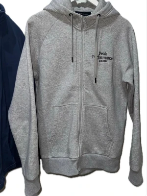 Grå zip hoodie från Peak Performance - Hej! Snygg gråmelerad hoodie från Peak Performance med dragkedja, huva med dragsko och två stora fickor framtill. Broderad logga på bröstet och mjukt, skönt material som passar perfekt till chill eller streetwear. Klassisk och enkel design. Den kostar 1099kr hos sportmore.com länk: https://sportamore.com/products/peak-performance-m-original-zip-hood-med-grey-melange. Jag säljer min för halva pris: 549kr 
