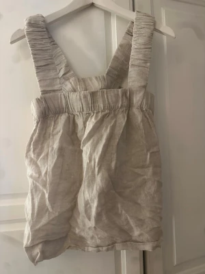 Beige linnetopp från kay/day - Säljer en beige linnetopp från kay/day med breda, rynkade axelband och elastisk kant upptill. Toppen är ärmlös och har en avslappnad passform, perfekt för varma dagar. Materialet känns luftigt och naturligt.