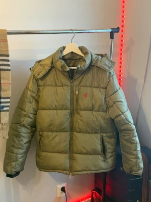 pufferjacka från Polo Ralph Lauren - Snygg olivgrön pufferjacka från Polo Ralph Lauren med dragkedja framtill, två sidofickor och ikonisk röd logga på bröstet. Jackan har hög krage och quiltad design som ger en riktigt schysst look. Perfekt för kalla vinterdagar.