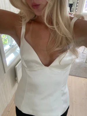 Vit satinlinnetopp med knytband - Snygg vit linnetopp i satin med djup v-ringning och breda axelband som knyts på axlarna. Toppen har en figurnära passform och slät yta som ger en elegant look. Perfekt att styla med jeans eller kjol för en trendig outfit.
