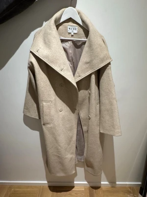 Beige oversized kappa från NA-KD - Stilren beige kappa från NA-KD med bred krage. Dubbelknäppt framtill, två fickor och slits bak. Tillverkad med helfodrad insida. Perfekt för lager på lager och en trendig look.