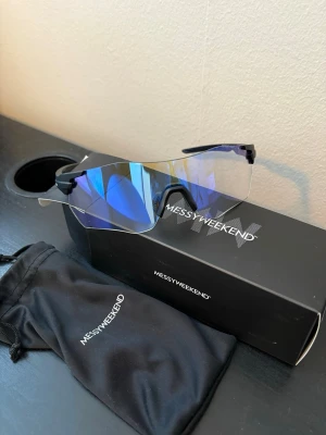 MessyWeekend Sonic – Sportiga solglasögon med färgskiftande lins - Säljer MessyWeekend Sonic – sportiga solglasögon med genomskinliga linser som skiftar färg efter ljus (från klar till blå/spegeleffekt). Cool, ramlös sköldformad design med svart båge som ger en futuristisk och trendig look.
