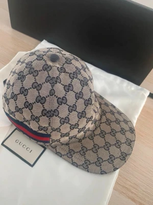 Gucci beige GG keps med röd-blå rand - Säljer en beige keps från Gucci med klassiskt GG-monogram i mörkblått över hela kepsen. På sidan finns en röd och blå rand som ger extra lyxig känsla. Justerbar rem bak i läder och insida i mörkblått tyg. Perfekt för dig som gillar exklusiva accessoarer.