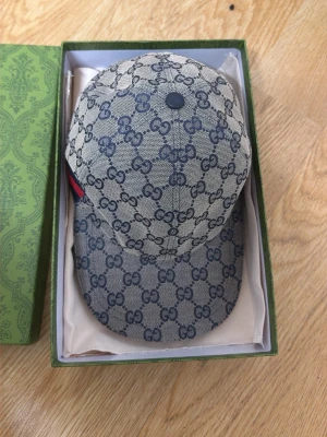 Gucci keps med GG-mönster - Säljer en snygg keps från Gucci med klassiskt GG-monogram i blått på beige botten. Kepsen har böjd skärm och justerbar baksida. Materialet är canvas och insidan är fodrad i mörkblått tyg. Perfekt för dig som gillar exklusiva accessoarer.