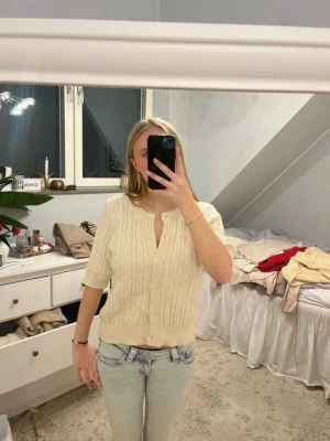 Kortärmad kabelstickad kofta i offwhite - Snygg kabelstickad kofta i offwhite med korta ärmar och rund hals. Koftan har knäppning framtill med matchande knappar och ribbade muddar vid ärmslut och nederkant. Perfekt att styla med jeans för en avslappnad look.