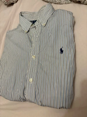 Randig skjorta från Polo Ralph Lauren - Klassisk vit skjorta med tunna blå ränder från Polo Ralph Lauren. Skjortan har knappar framtill, button-down krage och den ikoniska broderade loggan på bröstet. Tillverkad i bomull och har en snygg custom fit-passform.