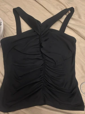 Svart linnetopp med twistade band - Snygg svart linnetopp med breda twistade axelband och rynkade detaljer framtill. Toppen är ärmlös och har en tight passform som framhäver figuren. Perfekt att styla med jeans eller kjol för en trendig look.