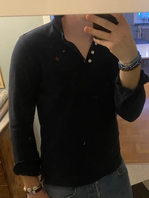 Mörkblå Rugbytröja från Morris - Snygg mörkblå Rugbytröja från Morris med klassisk krage. Skjortan har lång ärm och en liten broderad logga i röt på bröstet. Perfekt till jeans för en avslappnad men stilren look.