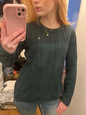 Mörkgrön kabelstickad tröja Bondelid - Mysig mörkgrön kabelstickad tröja från Bondelid med rund halsringning och diskret logga på bröstet. Tröjan har långa ärmar och ribbade muddar vid ärmslut och nederkant. Perfekt för lager på lager och chill höststil.