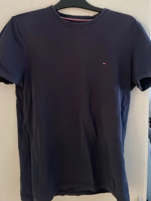 Mörkblå t-shirt från Tommy Hilfiger - Klassisk mörkblå t-shirt från Tommy Hilfiger med liten broderad logga på bröstet. T-shirten har rund halsringning och korta ärmar. Tillverkad i mjuk bomull för en skön känsla och enkel stil.