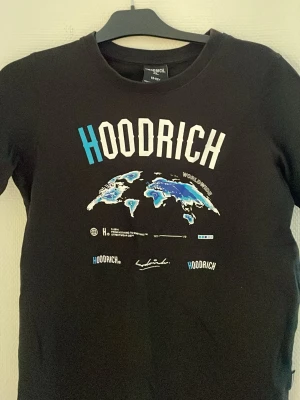 Svart HOODRICH t-shirt med världskarta - Cool svart t-shirt från Hoodrich med tryck av världskarta i blått och vitt samt stor HOODRICH-logga på bröstet. T-shirten har rund hals och korta ärmar, perfekt för en streetwear-look.