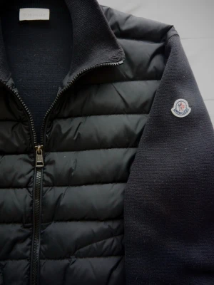 Svart Cardigan- Moncler - Svart quiltad cardiganjacka från Moncler med stickade ärmar och hög krage. Jackan har dragkedja framtill och Moncler-logga på ena ärmen. Perfekt för lager-på-lager och snygg till streetwear. NFC funkar såklart och allting ser ut som det ska! Sparsamt använd utan någon defekt.