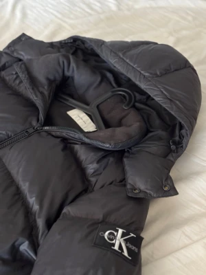 Svart jacka från Calvin Klein - Säljer en svart pufferjacka från Calvin Klein med huva och dragkedja framtill. Väldigt bra skick, inga hål, skador eller annat. Perfekt för kalla dagar och riktigt stilren design. Passar på mig som är 186cm lång men passar även till lite kortare