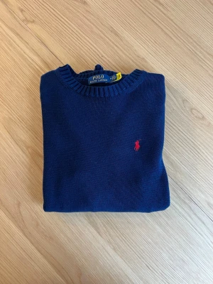 Mörkblå stickad tröja Polo Ralph Lauren - Klassisk mörkblå stickad tröja från Polo Ralph Lauren med rund halsringning och ribbade muddar. Tröjan har den ikoniska röda logotypen broderad på bröstet. Perfekt till jeans eller chinos för en clean och stilren look.