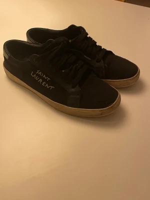 Svarta Saint Laurent sneakers - Säljer ett par svarta Saint Laurent sneakers med vit text på sidan. Skorna har lågt skaft, svarta skosnören och beige sula. Materialet är en mix av mocka och läder vilket ger en stilren och klassisk look. Det finns ett litet hål på sidan där plösen slutar men annars är dem väldigt fina. Box och dustbag ingår