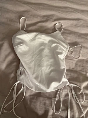 Vit linnetopp med snörning - Säljer en vit linnetopp med tunna axelband och snörning i sidorna. Toppen har en enkel, stilren design och är perfekt för varma dagar. Materialet är silkes. Passar till många olika outfits! Aldrig använd