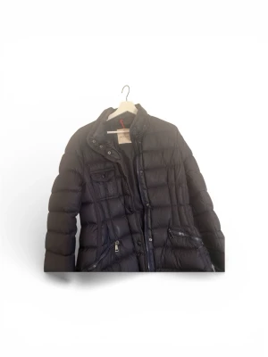 Svart dunjacka från Moncler - Snygg svart dunjacka från Moncler med quiltad design, hög krage och flera fickor med dragkedja. Insidan har det klassiska Moncler-serietrycket. Perfekt för kalla vinterdagar och riktigt stilren look.