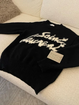 Svart stickad Saint Laurent-tröja - Säljer en svart stickad tröja från Saint Laurent med vit, grafisk logga framtill. Tröjan har rund halsringning och ribbade muddar vid ärmslut och nederkant. Materialet ser mjukt och fluffigt ut, perfekt för dig som gillar exklusiva streetwear-vibbar.