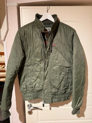 Olivgrön vindjacka från G-Star Raw - Snygg olivgrön vindjacka från G-Star Raw med quiltat foder i orange. Jackan har hög krage, dragkedja framtill, två stora fickor och ribbade muddar vid ärmslut och midja. Perfekt för dig som vill ha en stilren och funktionell jacka i slitstarkt material.