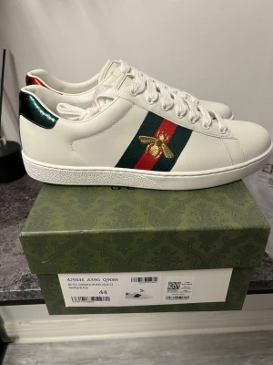 Gucci Ace vita sneakers med bi - Gucci Ace sneakers i vitt läder med klassiska gröna och röda sidoremsor och broderad guldfärgad bi. Metallicgrön detalj på hälen och vita skosnören. Snygg och tidlös design som sticker ut med exklusiva detaljer.