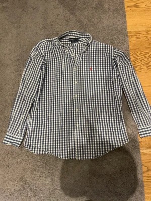 Rutig skjorta från Ralph Lauren - Klassisk långärmad skjorta från Ralph Lauren i blått och vitt rutigt mönster. Skjortan har knappar framtill och den ikoniska röda broderade loggan på bröstet. Tillverkad i mjuk bomull och har en avslappnad passform.
