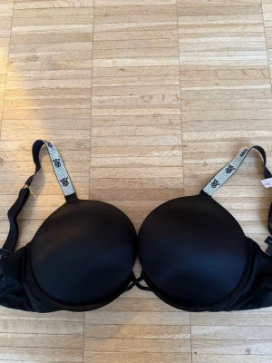 Svart push-up bh Victoria's Secret - Snygg svart push-up bh från Victoria's Secret med vadderade kupor och justerbara axelband dekorerade med glittrande stenar och VS-logga. Helt oanvänd i storlek 34 DD vilket motsvarar  75E/75DD