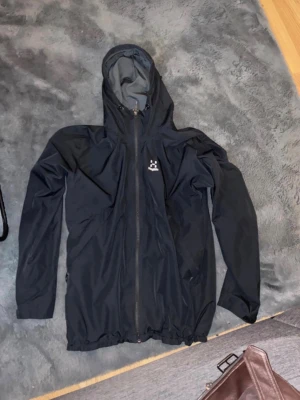 Svart regnjacka från Haglöfs - Snygg svart regnjacka från Haglöfs med hel dragkedja framtill och justerbar huva. Jackan har minimalistisk design, diskret logga på bröstet och är tillverkad i ett lätt, vattenavvisande material. Perfekt för regniga dagar och outdoor-äventyr.