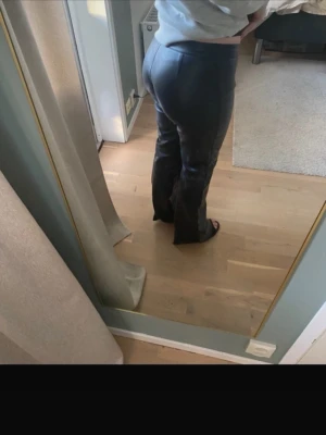 Svarta skinnbyxor med utsvängda ben - Säljer ett par svarta skinnbyxor med hög midja och utsvängda ben. Byxorna har en clean look utan synliga detaljer och stängs med knapp och dragkedja framtill. Perfekta för dig som vill ha en edgy och trendig stil.