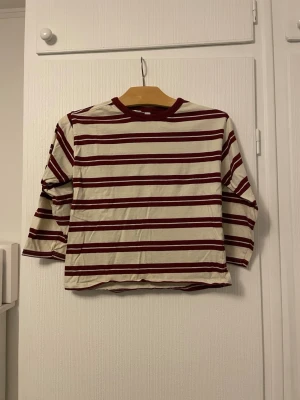 Randig topp med vinröda detaljer - Säljer en vit topp med vinröda horisontella ränder och matchande vinröd kant vid halsen. Toppen har trekvartsärm och en rak, något croppad passform. Perfekt för en avslappnad och trendig look.