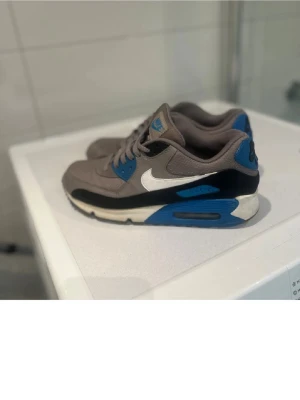 Nike Air Max 90 grå/blå sneakers - Nike Air Max 90 sneakers i grått, svart och blått med vit swoosh. Klassisk siluett med synlig Air-enhet i sulan och detaljer i mocka och mesh. Perfekta för dig som gillar streetwear och sportig stil.
