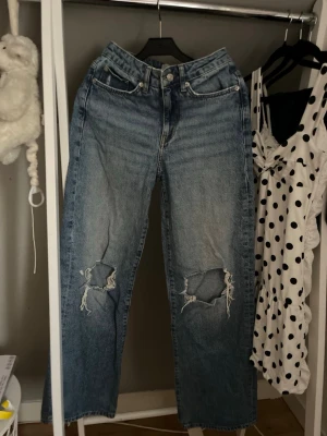 Blå slitna raka jeans med hål - Säljer ett par blå raka jeans med slitningar och stora hål på båda knäna. Jeansen har klassisk femficksmodell, hög midja och är gjorda i denim. Perfekta för en avslappnad och trendig look.