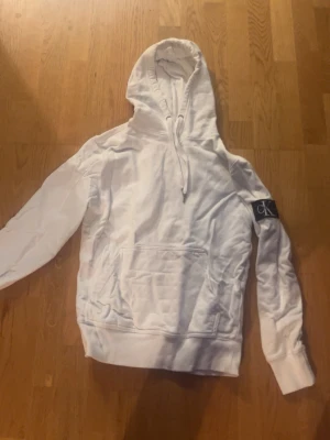 Vit hoodie med CK-logga - Säljer en vit hoodie från Calvin Klein med stor huva och dragsko. Klassisk känguruficka framtill och en svart CK-logga på vänster ärm. Tillverkad i mjukt bomullsmaterial och har ribbade muddar vid ärmslut och nederkant.