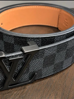 Svart och grått rutigt LV-bälte - Snyggt Louis Vuitton-bälte i svart och grått med klassiskt rutigt mönster och stor LV-logga i metall framtill. Bältet är gjort i skinn med en lyxig känsla och passar perfekt till jeans eller kostymbyxor.