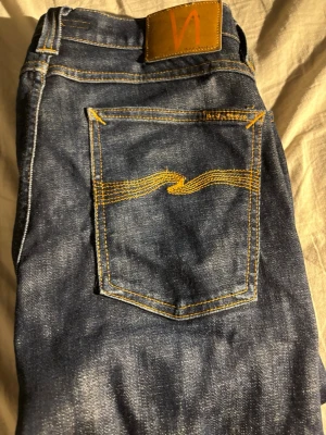 Mörkblå jeans från Nudie Jeans - Säljer ett par mörkblå jeans från Nudie Jeans med klassisk gul söm på bakfickan och läderpatch i midjan. Modellen är slim fit med raka ben och tillverkad i denim. Perfekta för dig som gillar stilrena och tidlösa jeans. Säljes pga av för långa!