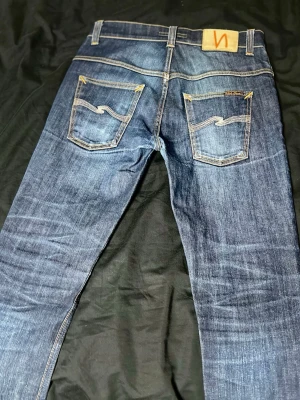 Blå jeans från Nudie Jeans - Säljer ett par klassiska blå jeans från Nudie Jeans med snygga slitningar och kontrastsömmar. Jeansen har raka ben, två bakfickor med den ikoniska Nudie-bågen och patch i läder bak i midjan. Tillverkade i slitstark denim, perfekta för en avslappnad stil.