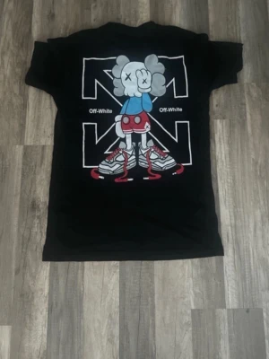 Ny Off-White t-shirt - Säljer en ny svart t-shirt från Off-White storlek M med stort grafiskt tryck på ryggen och mindre tryck framtill. Motivet är en tecknad figur med sneakers och färgglada detaljer i blått, rött och grått. Klassisk passform och rund hals. Perfekt för dig som gillar streetwear och coola prints. 