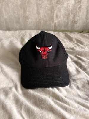 Svart Chicago Bulls keps - Säljer en svart keps med Chicago Bulls logga broderad framtill i rött och vitt samt texten 'BULLS' på sidan. Klassisk böjd skärm och justerbar passform. Tillverkad i bomullstyg, perfekt för dig som gillar sportig streetstyle.