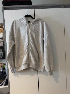 Ljusgrå zip hoodie från Gina Tricot - Ljusgrå hoodie med dragkedja från Gina Tricot. Klassisk modell med huva och dragsko, ribbade muddar vid ärmslut och nederkant samt två fickor framtill. Perfekt för en chill och avslappnad stil.
