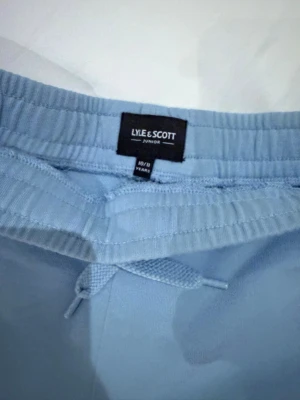 Ljusblå shorts från Lyle & Scott - Säljer ett par ljusblå shorts från Lyle & Scott med elastisk midja och snörning framtill. Shortsen har en klassisk passform och är tillverkade i mjuk bomull. På vänster ben finns den ikoniska gula loggan broderad.