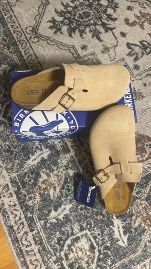 Beige Birkenstock Boston tofflor - Snygga beige Birkenstock Boston tofflor i mocka med justerbart spänne i metall. Klassisk slip-in modell med mjuk fotbädd och rundad tå. Perfekta för en chill och stilren look hemma eller ute. Levereras med originalkartong och tagg.