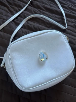Vintage Caroline Svedbom Axelväska Vit - Snygg vit vintage axelväska i fyrkantig modell med lång justerbar rem. Väskan har en stor ”mjölkvit” glittrande kristall på framsidan som ger en unik och ikonisk look. Tillverkad i slätt konstläder och stängs med dragkedja. Väldigt sällsynt väska då den är köpt många år sedan och ej görs längre. Perfekt för dig som vill sticka ut. 