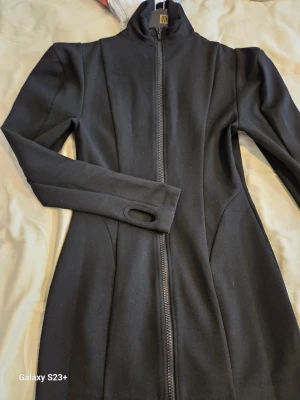 Svart klänning med dragkedja och tumhål - Snygg svart klänning med hög krage och hel dragkedja framtill. Klänning har långa ärmar med tumhål och en figurnära passform. Perfekt för dig som gillar en stilren och sportig look.