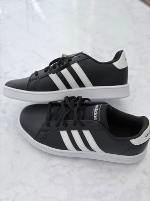 Adidas Grand Court svartvita sneakers- barn strl 31/2 - Snygga Adidas Grand Court sneakers i svart med klassiska vita ränder på sidorna och vit sula. Skorna har lågt skaft, snörning och är tillverkade i syntetmaterial. Perfekta för en sportig och stilren look.storlek 31/2