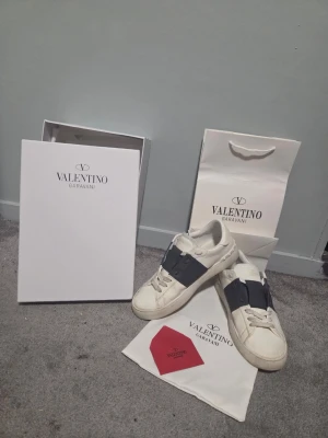 Valentino Garavani Open - Snygga vita sneakers från Valentino Garavani med bred navy blue rem över vristen och gulddetaljer på plösen. Skorna har klassisk låg profil och är tillverkade i läder med vita snören. Perfekta för dig som vill ha en stilren och exklusiv look. Litet hål som syns på den sista bilden.