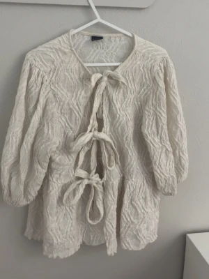 Krämvit blus med knytband - Trendig krämvit blus från Gina tricot  i strukturerat tyg med tre dekorativa knytband framtill. Blusen har en lös passform och trekvartsärmar med resår vid ärmsluten. Perfekt för en avslappnad men stilren look.😍
