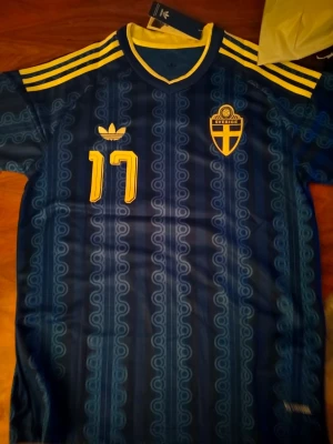 Blå Sverige fotbollströja Adidas - Säljer en blå fotbollströja från Adidas med Sveriges landslagsemblem och nummer 17 i gult. Tröjan har gula detaljer på axlarna, mönstrad design och namnet Gyökeres på ryggen. Perfekt för dig som vill stötta Sverige på match!