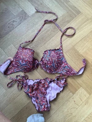 Mönstrad bikini med snörning - Snygg bikini med paisleymönster i rosa, lila och röda toner. Överdelen har vadderade kupor och knytband i nacken, medan underdelen har snörning i sidorna. Perfekt för sommaren och strandhäng!