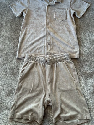 Beige loungewear set med shorts - Mysigt loungewear set i beige frotté med kortärmad skjorta och matchande shorts. Skjortan har knappar framtill och bröstficka, shortsen har resår i midjan, sidofickor och en bakficka. Perfekt för chill dagar hemma.