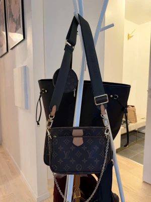 Brun monogram crossbody handväska - Snygg brun handväska med klassiskt monogrammönster och detaljer i beige och guld. Väskan har bred axelrem i textil, kedjedetalj och en liten rund myntväska. Perfekt för dig som gillar trendiga accessoarer och vill ha plats för det viktigaste.